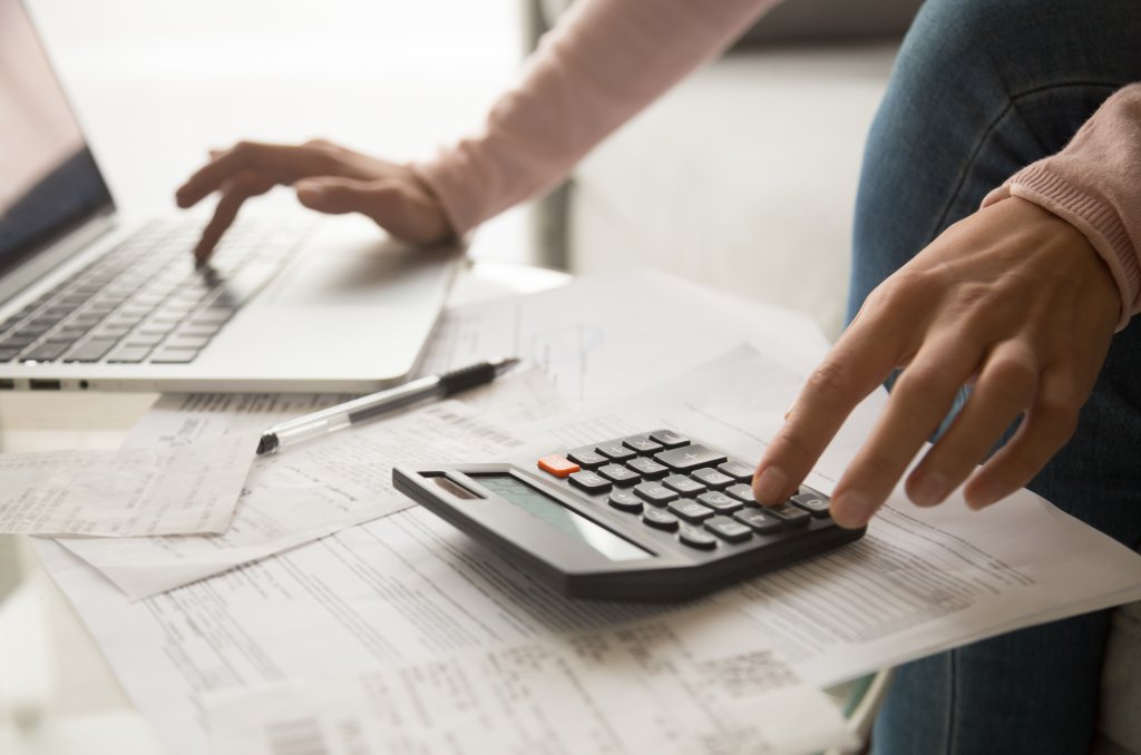 Close up cropped image young woman calculating monthly expenses, managing budget, entering data in computer application, sitting at table full of papers, loan documents, invoices, utility bills. Close up cropped image young woman calculating monthly expenses, managing budget, entering data in computer application, sitting at table full of papers, loan documents, invoices, utility bills.
